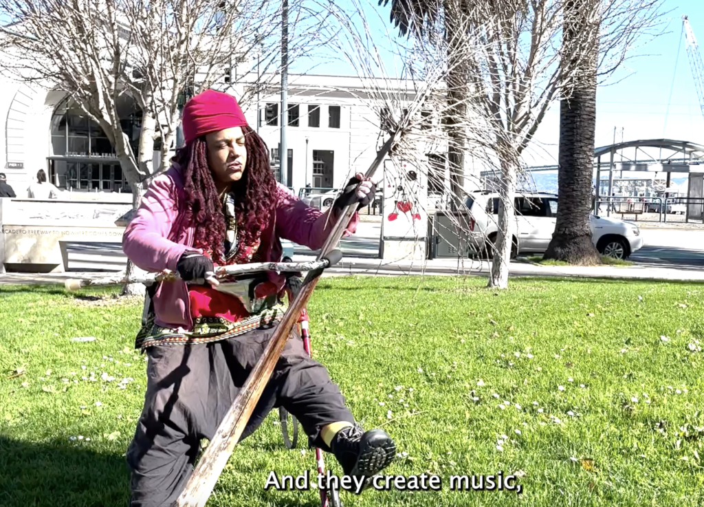 Claudia Alick, a black queer person with a walker and a cane makes music with a dried palm frond at the Embarcadero in San Francisco. Claudia Alick, a black queer person with a walker and a cane makes music with a dried palm frond at the Embarcadero in San Francisco.