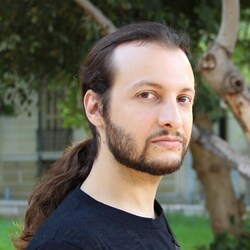 Portrait of Panas Karampampas, a persom with long dark hair tied back in a ponytail and a short beard, wearing a black T-shirt, standing outdoors with trees and greenery in the background.