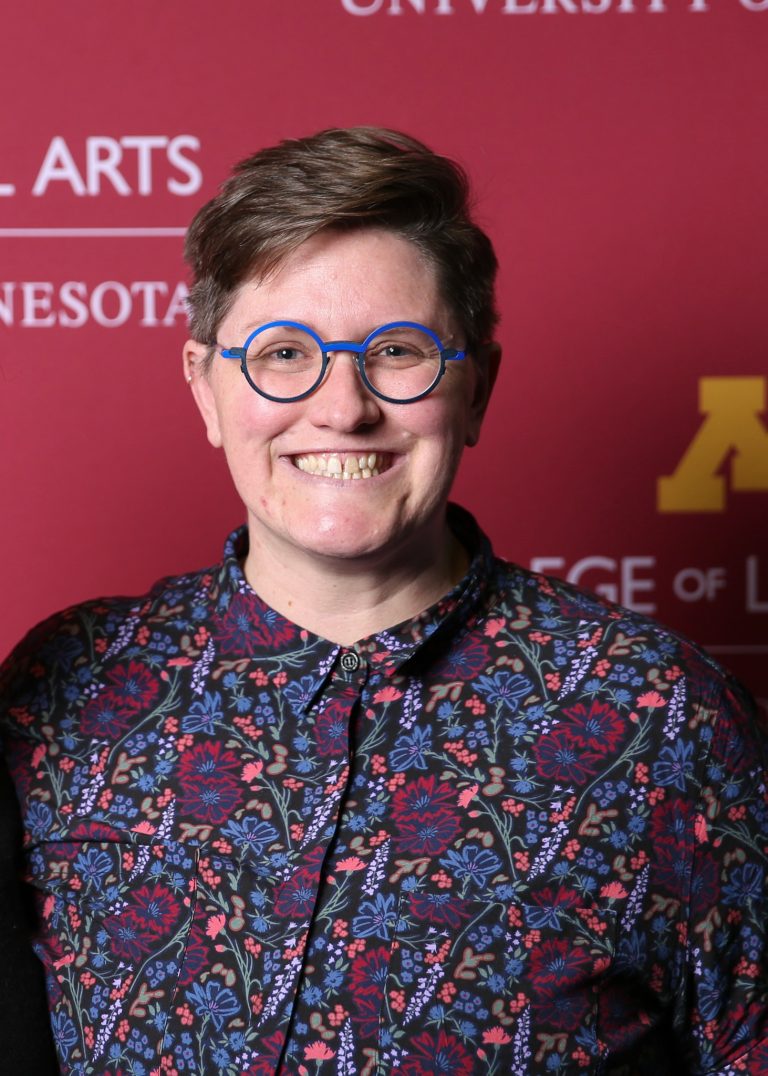 Photo of a smiling white, nonbinary professor with short brown hair, whimsical blue glasses, and a flowered button up shirt